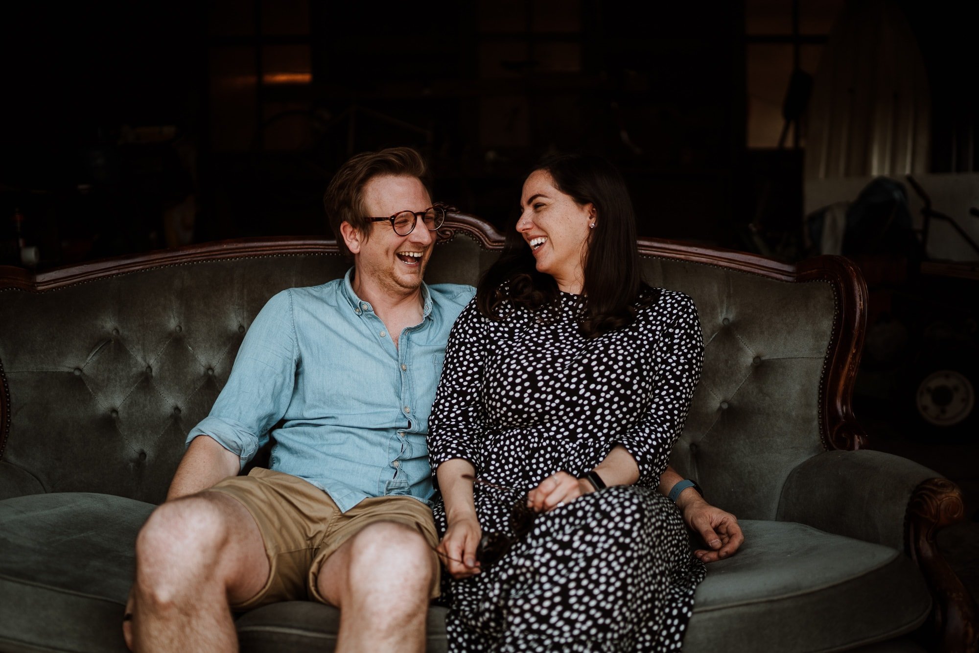 Roxanne and Ed from The Little Wedding Warehouse seated relaxed and laughing on grey sofa in rustic barn during their branding photoshoot