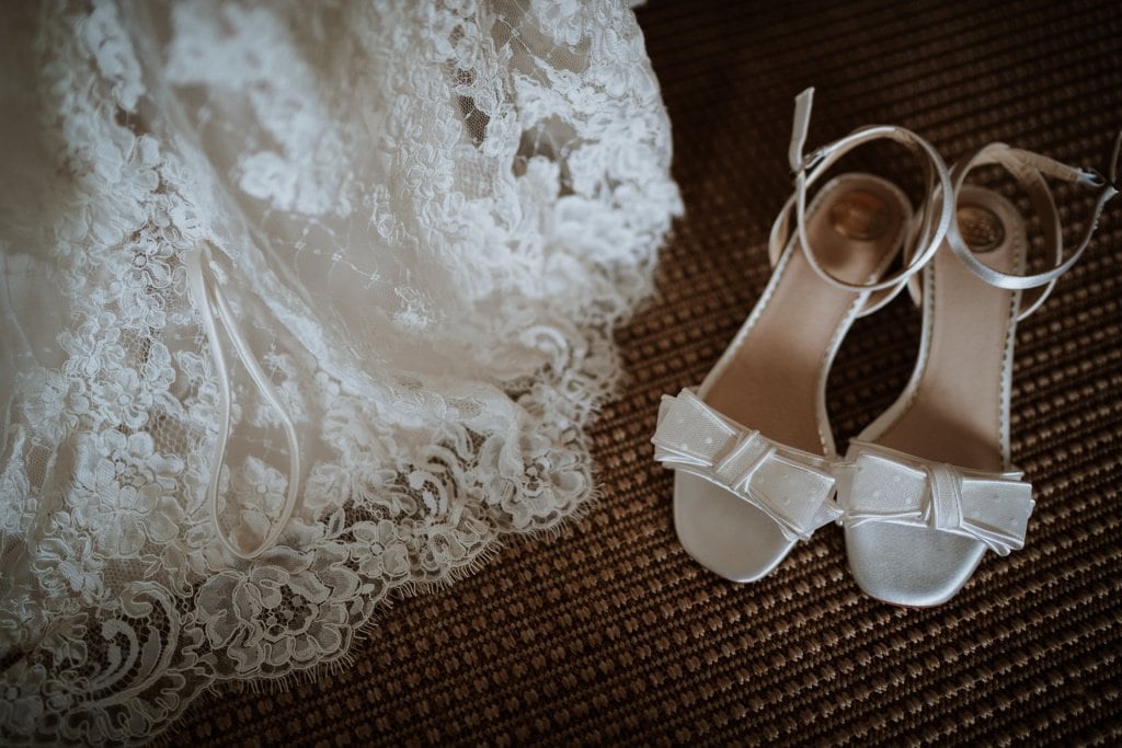 Beautiful bridal sandals photographed next to the bottom of a stunning lace wedding dress