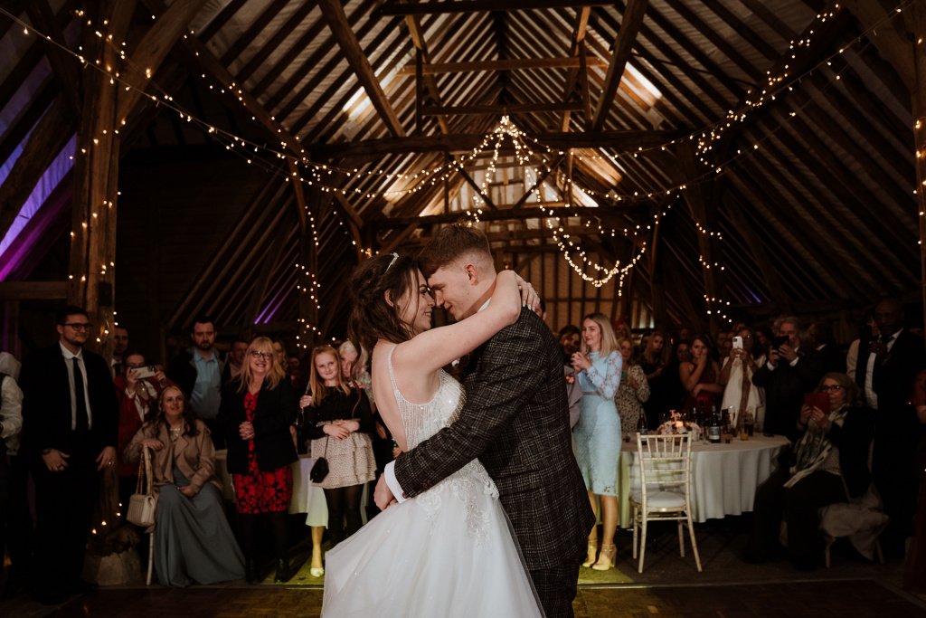 Bride and Groom cuddling each other during their first dance, beneath fairy lights and watched by their guests
