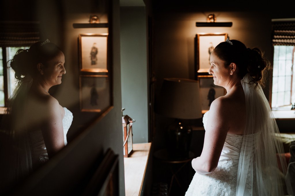 Brunette Bride stands in front of the window and her reflection in a picture frame can be seen