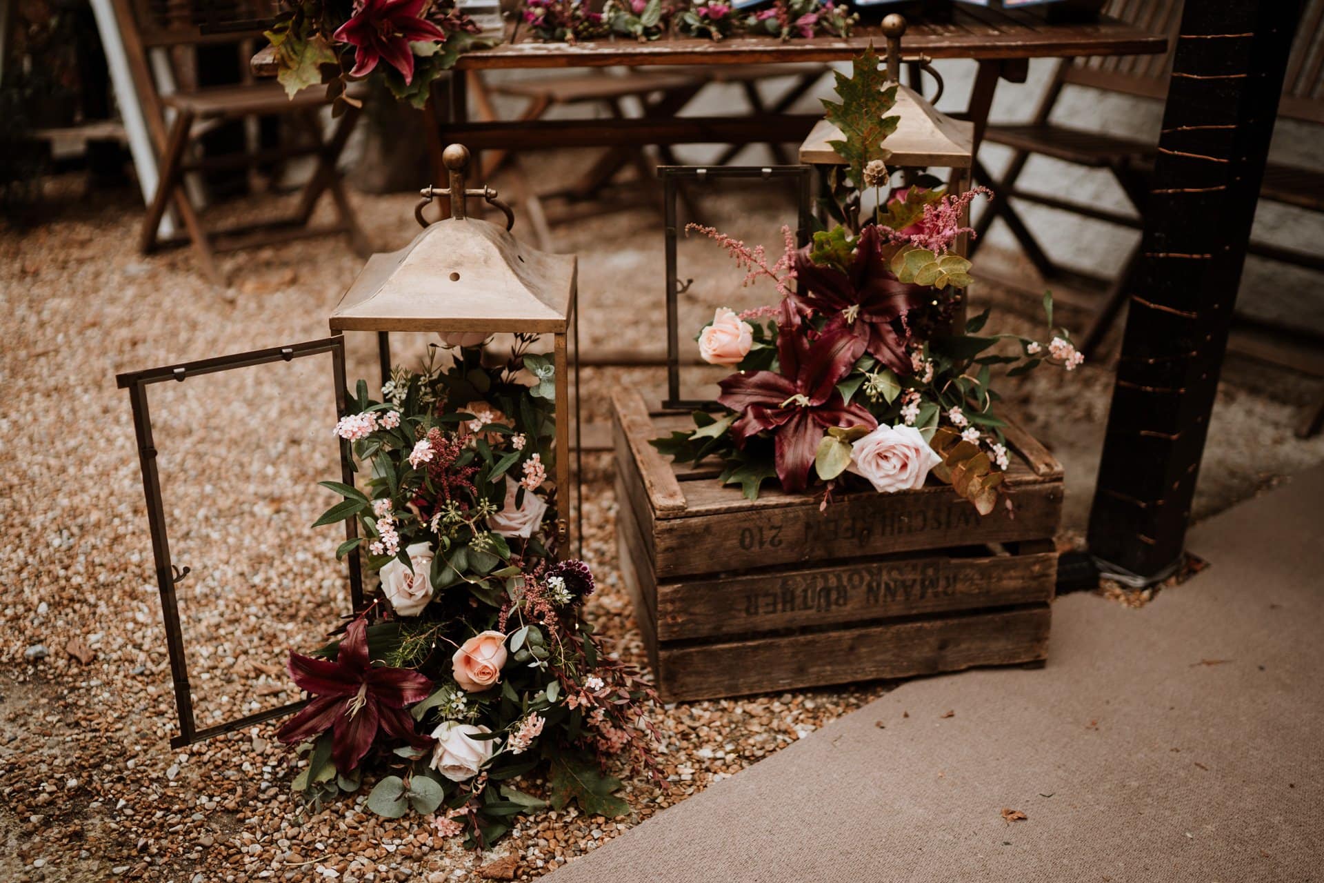 Glass lanterns with the door open with burgundy and blush pink flowers overflowing out next to rustic wooden crate adorned with more flowers