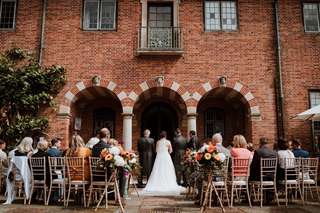 Wedding ceremony taking place set against red brick wall arches and bright exotic flowers lining the aisle