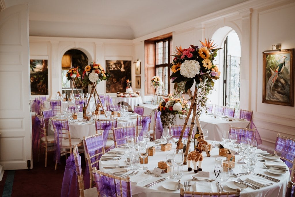 The wedding breakfast room of Port Lympne Hotel dressed for a wedding with bright floral displays on the tables and purple chair sashes