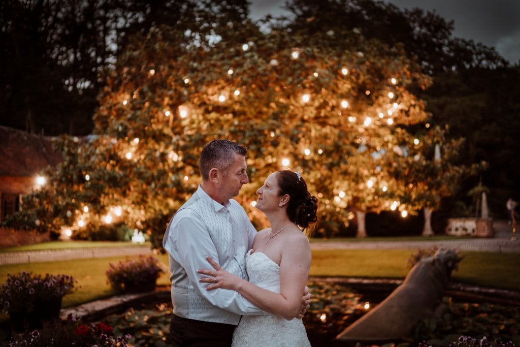 Bride and Groom cuddling infront of a large tree illuminated with festoon lighting
