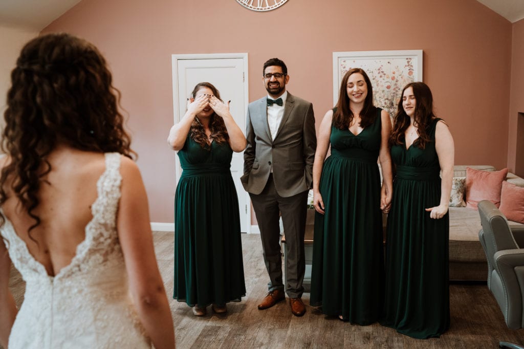 Bride standing in front of her bridesmaid and brides man who have their eyes closed waiting to see her for the first time
