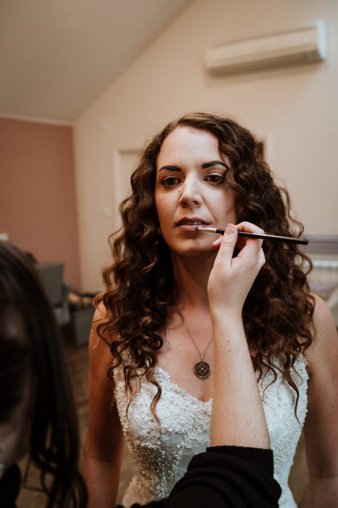 Bride with long dark wavy hair having her lip colour applied before her wedding ceremony
