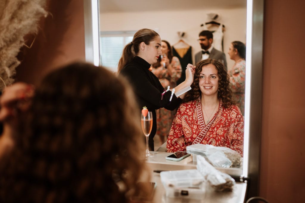 Bride to be wearing a red robe looking in a mirror having her long dark curly hair done for her wedding day