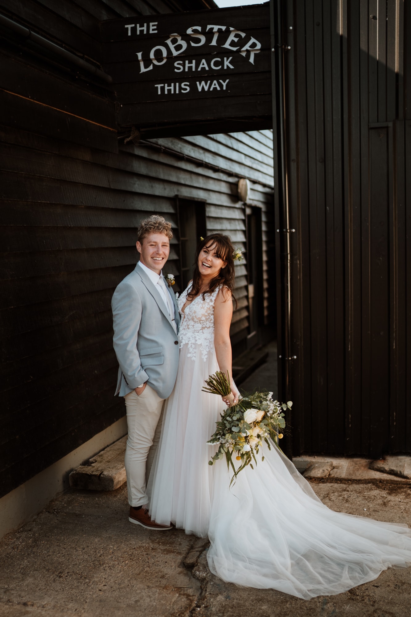 Stunning bride and groom laughing and looking relaxed under a sign for the lobster shack at East Quay wedding venue