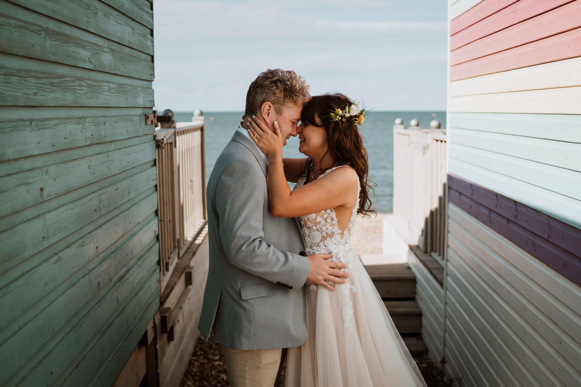 Bride holding grooms face as they kiss between beach huts at East Quay, Kent wedding venue