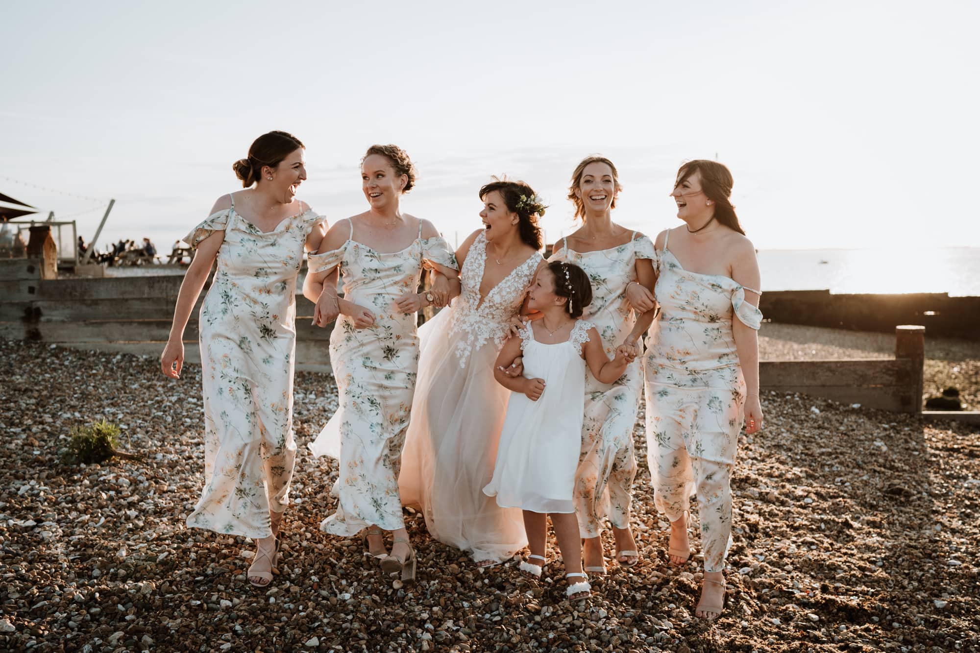 Bride and her bridesmaids laughing together arm in arm as they walk along Whitstable beach